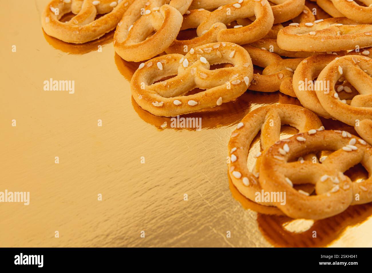 I deliziosi pretzel dalla consistenza croccante e cosparsi di semi di sesamo poggiano su una superficie dorata riflettente, perfetti per spuntini o incontri Foto Stock