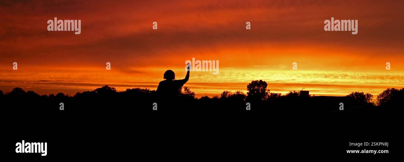 Tramonto su campi, orizzonte lontano, alberi ed edifici sagomati contro un cielo colorato. Persona che saluta la buonanotte al sole. Foto Stock