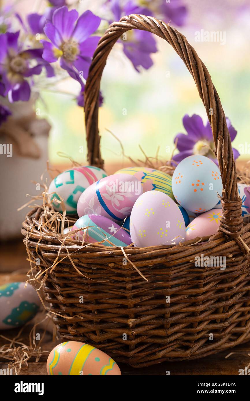 Uova di Pasqua in un cestino e margherite viola su un tavolo di legno con colorato sfondo primaverile Foto Stock