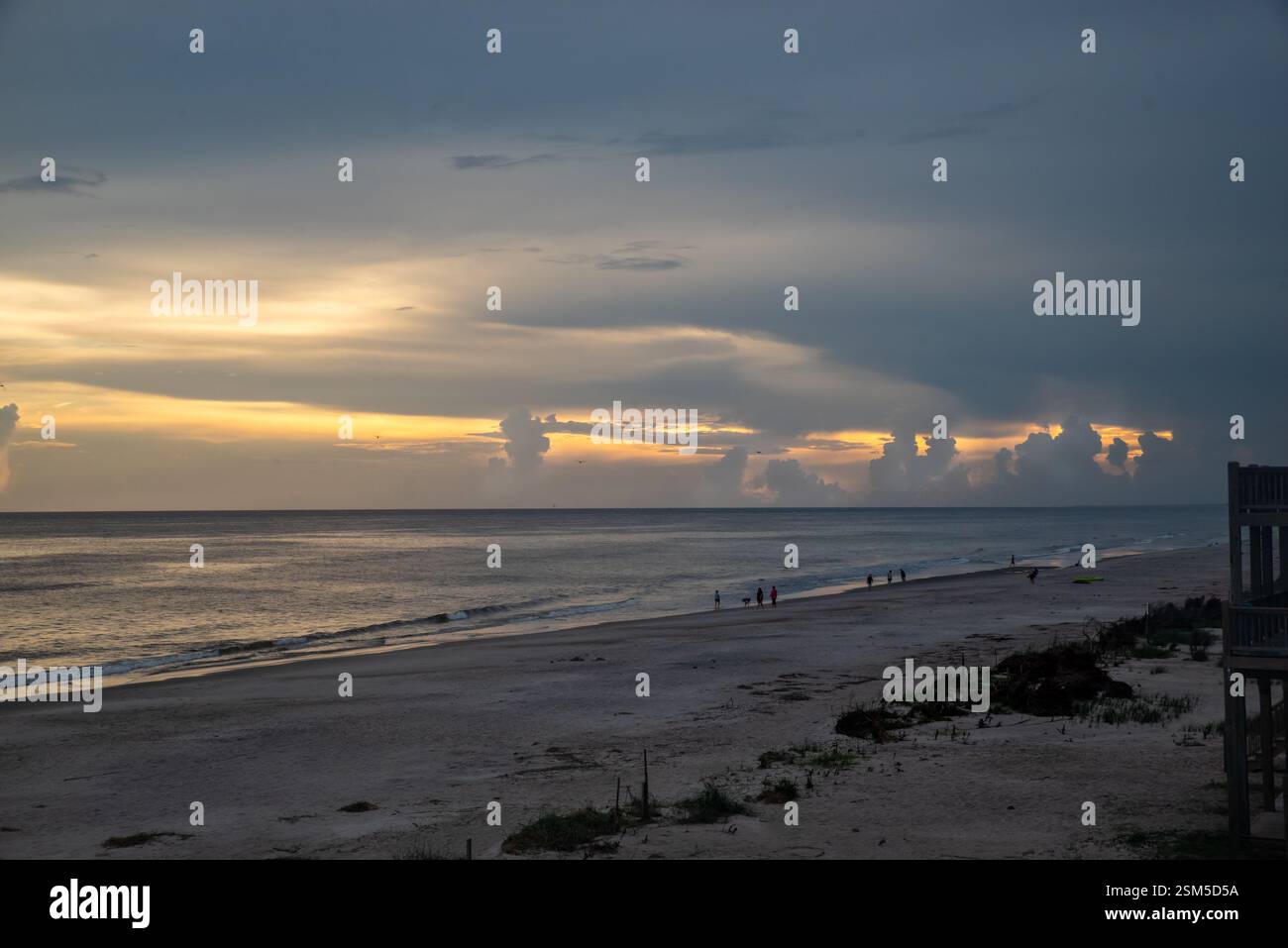 Una spiaggia tranquilla al tramonto con una luce dorata che attraversa le nuvole sull'oceano mentre le onde dolci incontrano la riva e la gente cammina lungo la costa. Foto Stock