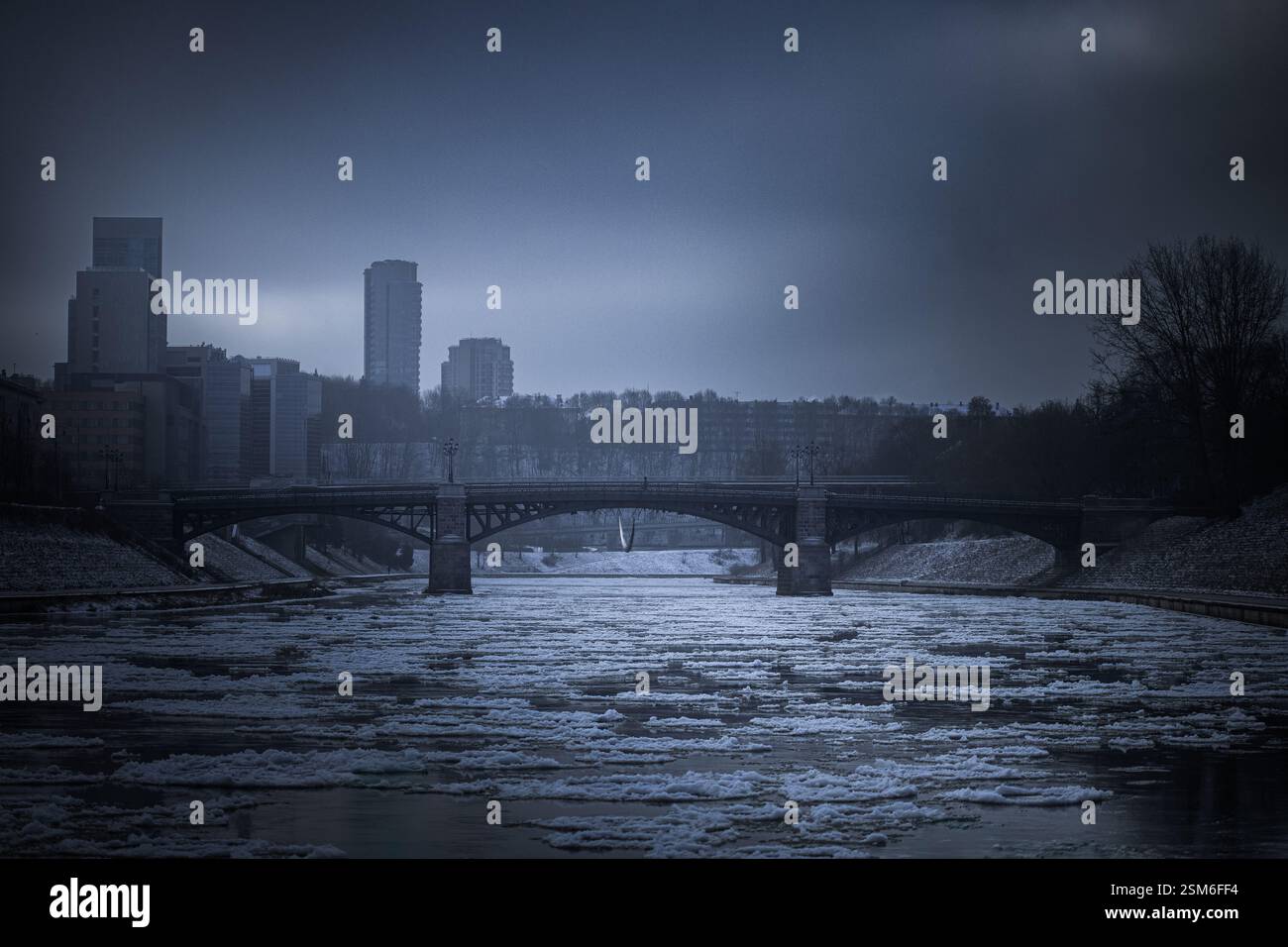 Una fredda scena invernale di un ponte su un fiume parzialmente ghiacciato a Vilnius, Lituania. L'atmosfera scura e soffusa aggiunge un tocco cinematografico a questa città urbana Foto Stock