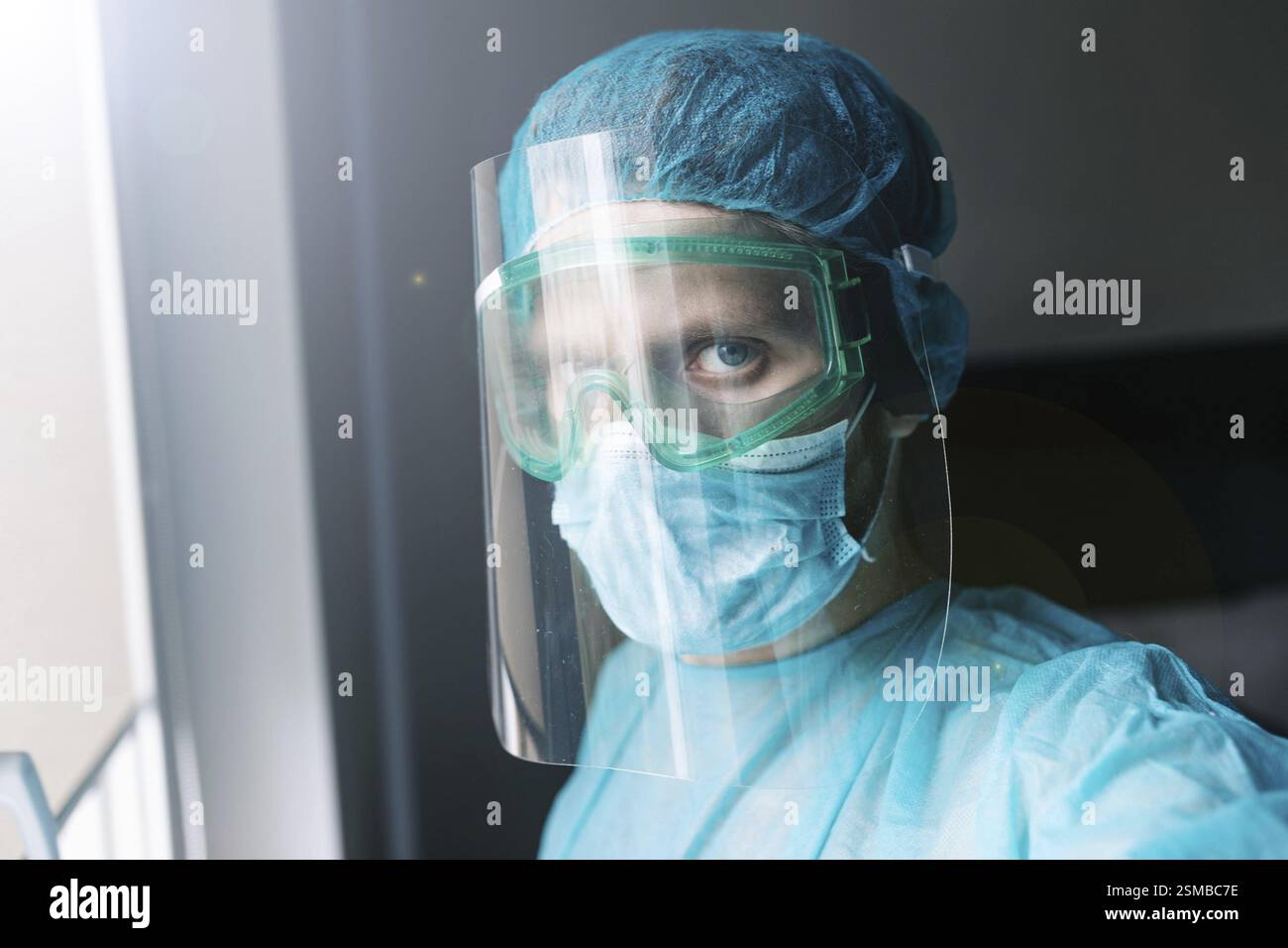Operatore medico in abbigliamento protettivo blu e schermo viso vicino a una finestra, apparendo vigile Foto Stock