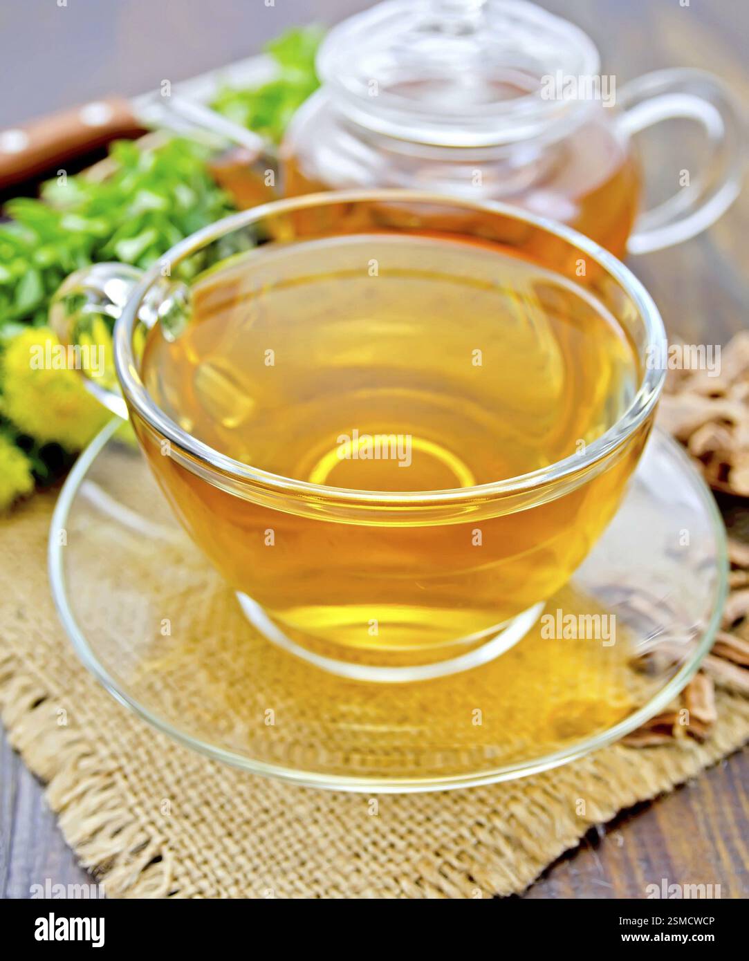 Il tè alle erbe in un bicchiere di vetro, una teiera su un tovagliolo di tela ruvida, radice secca e fiori freschi Rhodiola rosea sullo sfondo delle tavole di legno Foto Stock