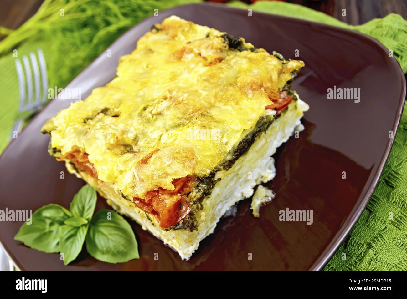 La torta di patate e formaggio, pomodoro e spinaci, uova riempite con il latte in una piastra marrone su un tovagliolo al buio su un pannello di legno Foto Stock
