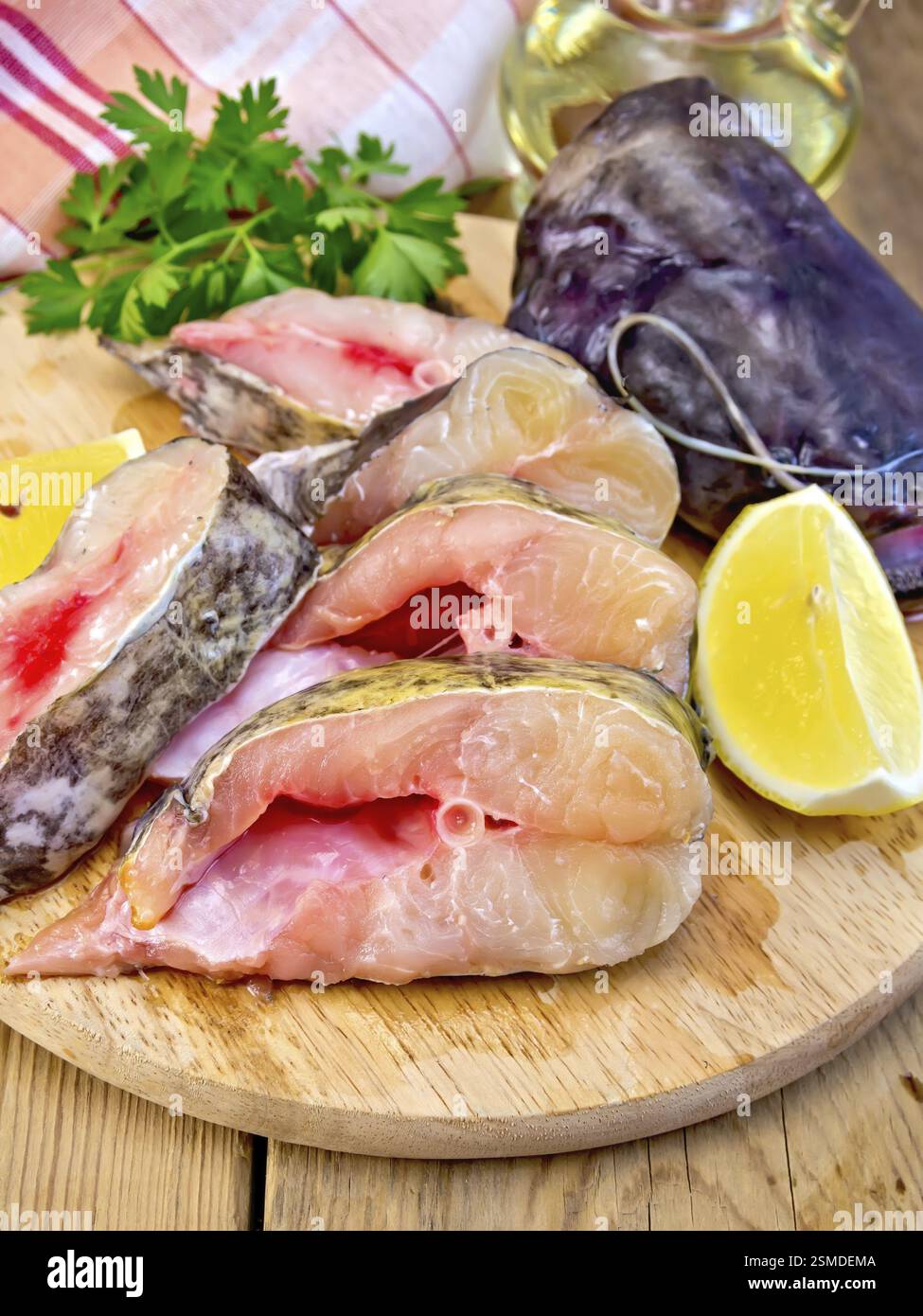 Pezzetti di pesce gatto a fette crude su un bordo circolare, prezzemolo, limone, olio, panno su uno sfondo di tavole di legno Foto Stock