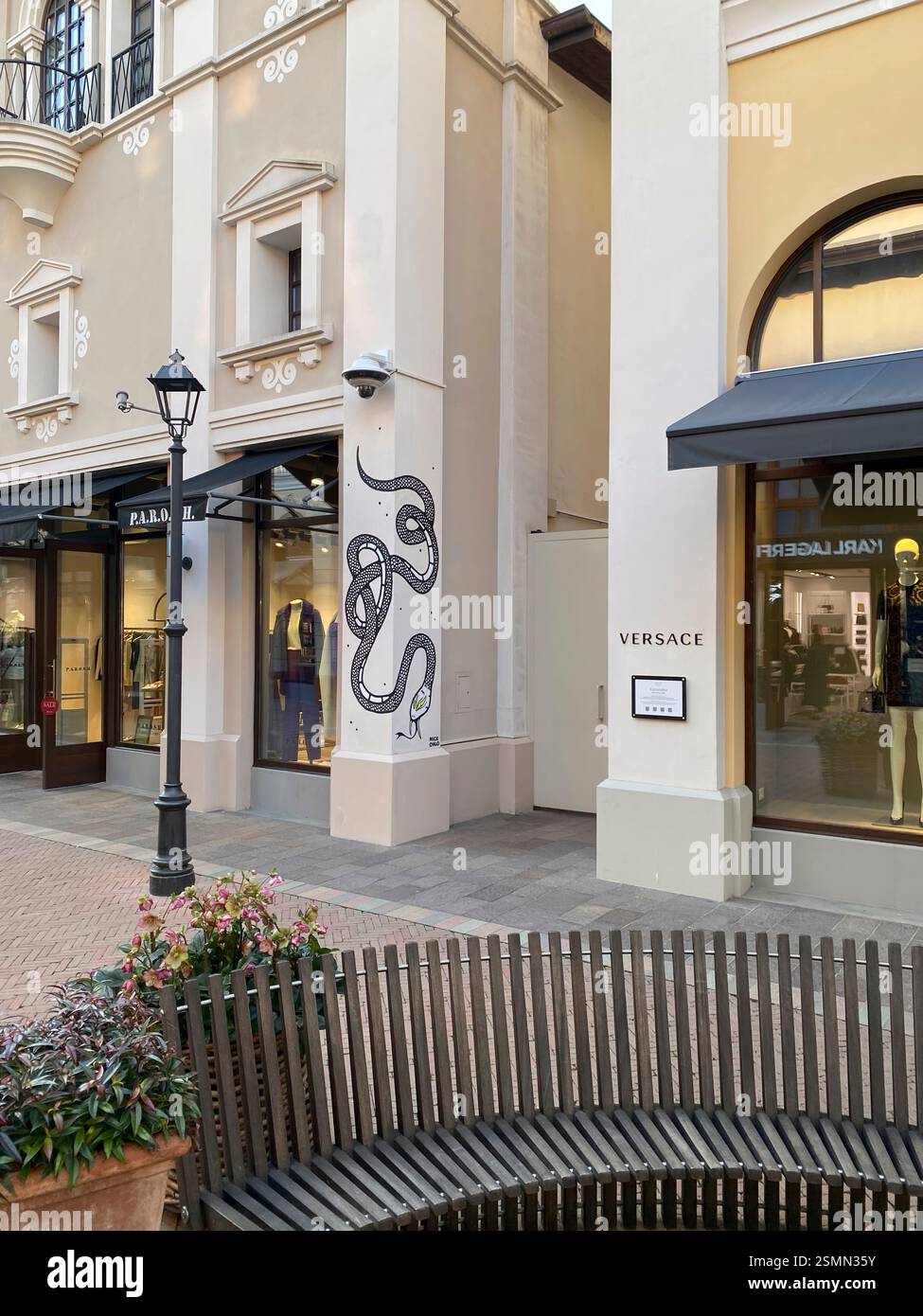 Panca vuota di fronte ai negozi di stilisti del centro commerciale outlet di lusso Fidenza Village in Italia, con cartellonistica per Street art e Versace Foto Stock
