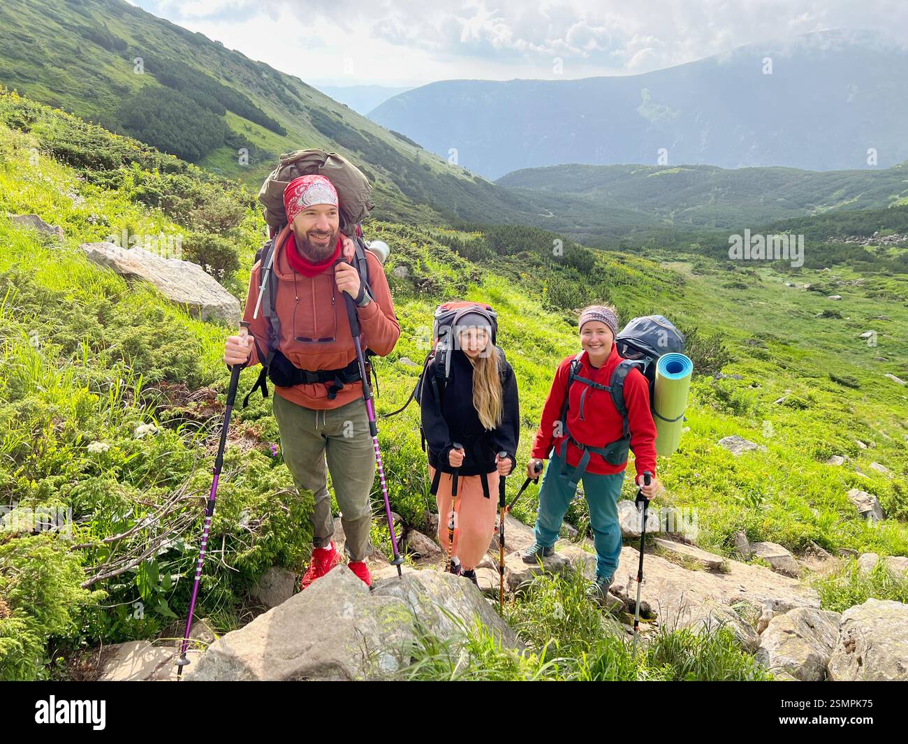 Tre escursionisti, vestiti con colorate attrezzature all'aperto, fanno una pausa su un sentiero roccioso e verde. Lo sfondo panoramico presenta colline lussureggianti e un cielo parzialmente nuvoloso. Foto Stock