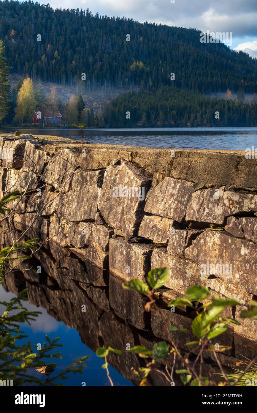 Tranquilla estate a Lunner Almenning: Laghi tranquilli, una diga di pietra con acqua corrente e una barca solitaria che si sposta nel tranquillo abbraccio della natura. Un retre pacifico Foto Stock