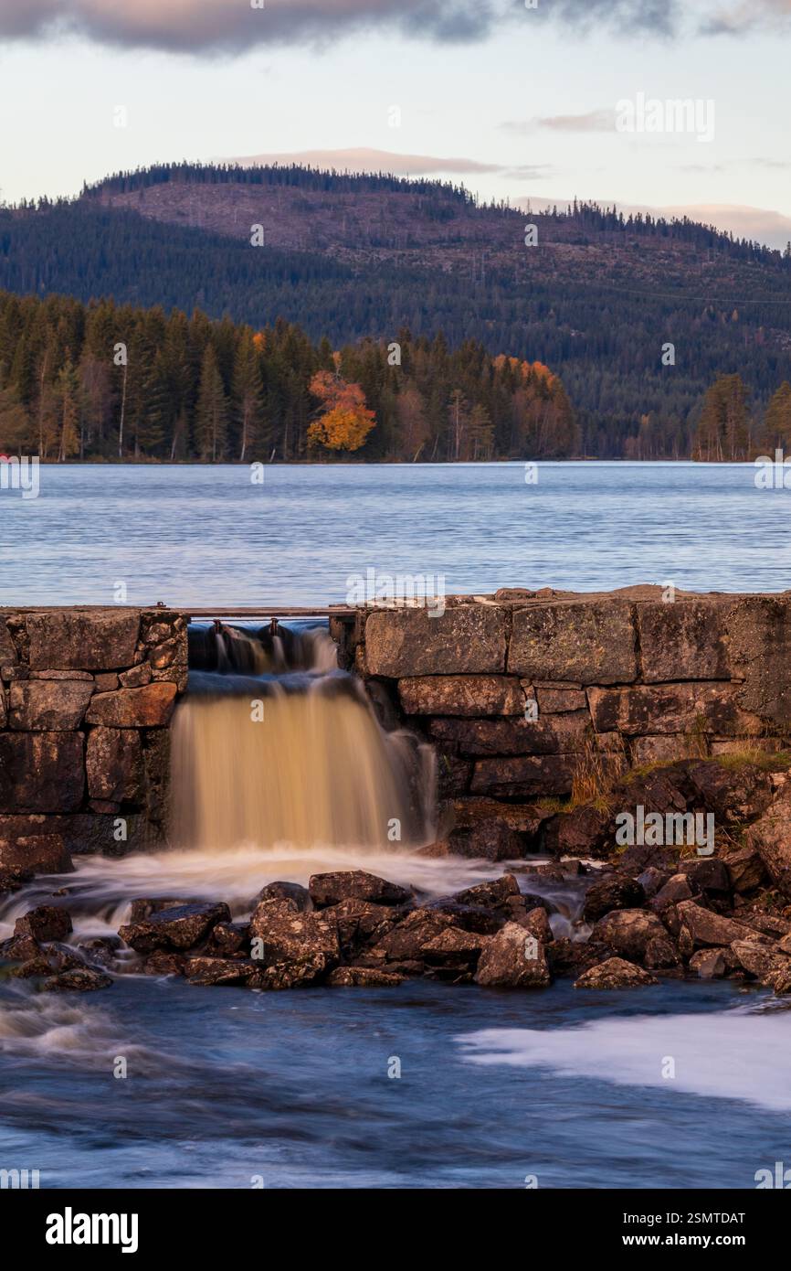 Tranquilla estate a Lunner Almenning: Laghi tranquilli, una diga di pietra con acqua corrente e una barca solitaria che si sposta nel tranquillo abbraccio della natura. Un retre pacifico Foto Stock