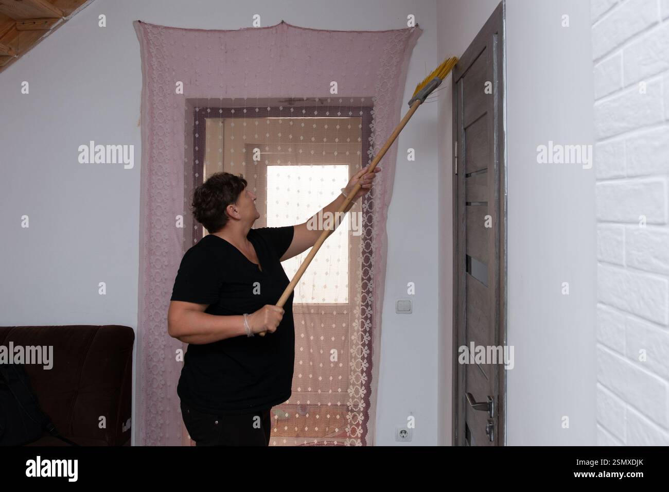 Si vede una donna che pulisce la parte superiore di un telaio della porta con un panno a manico lungo. Si trova in una stanza parzialmente arredata, con riccio decorativo puro Foto Stock