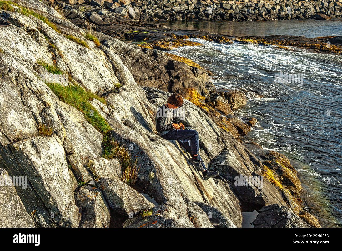 Giovane seduto e rilassato sulle rocce della Atlantic Road in Norvegia, godendosi il panorama. Ponte Hulvågen, Vevang, Norvegia, Europa Foto Stock