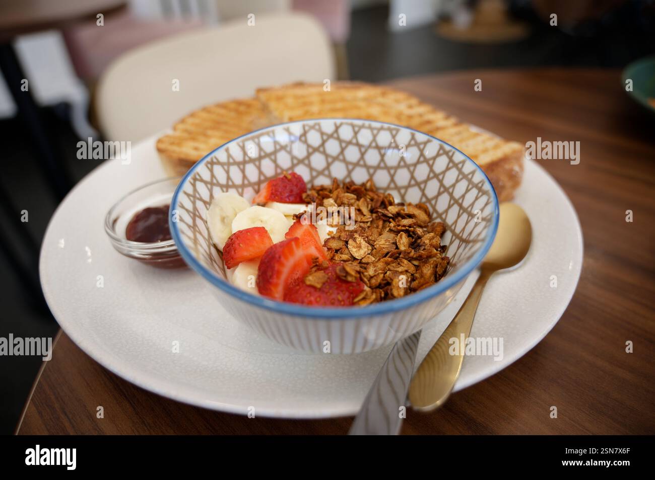 Una sana ciotola per colazione con yogurt, muesli, fragole fresche e fette di banana, servita con pane tostato e marmellata Foto Stock
