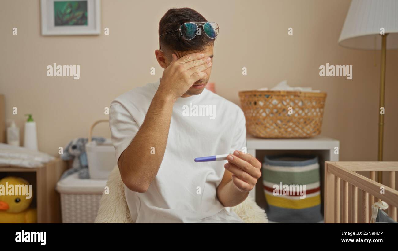 Giovane ispanico nella stanza del bambino che tiene il test di gravidanza con la mano sulla fronte, circondato da culla e oggetti per bambini, trasmettendo emozioni e anticipazione Foto Stock