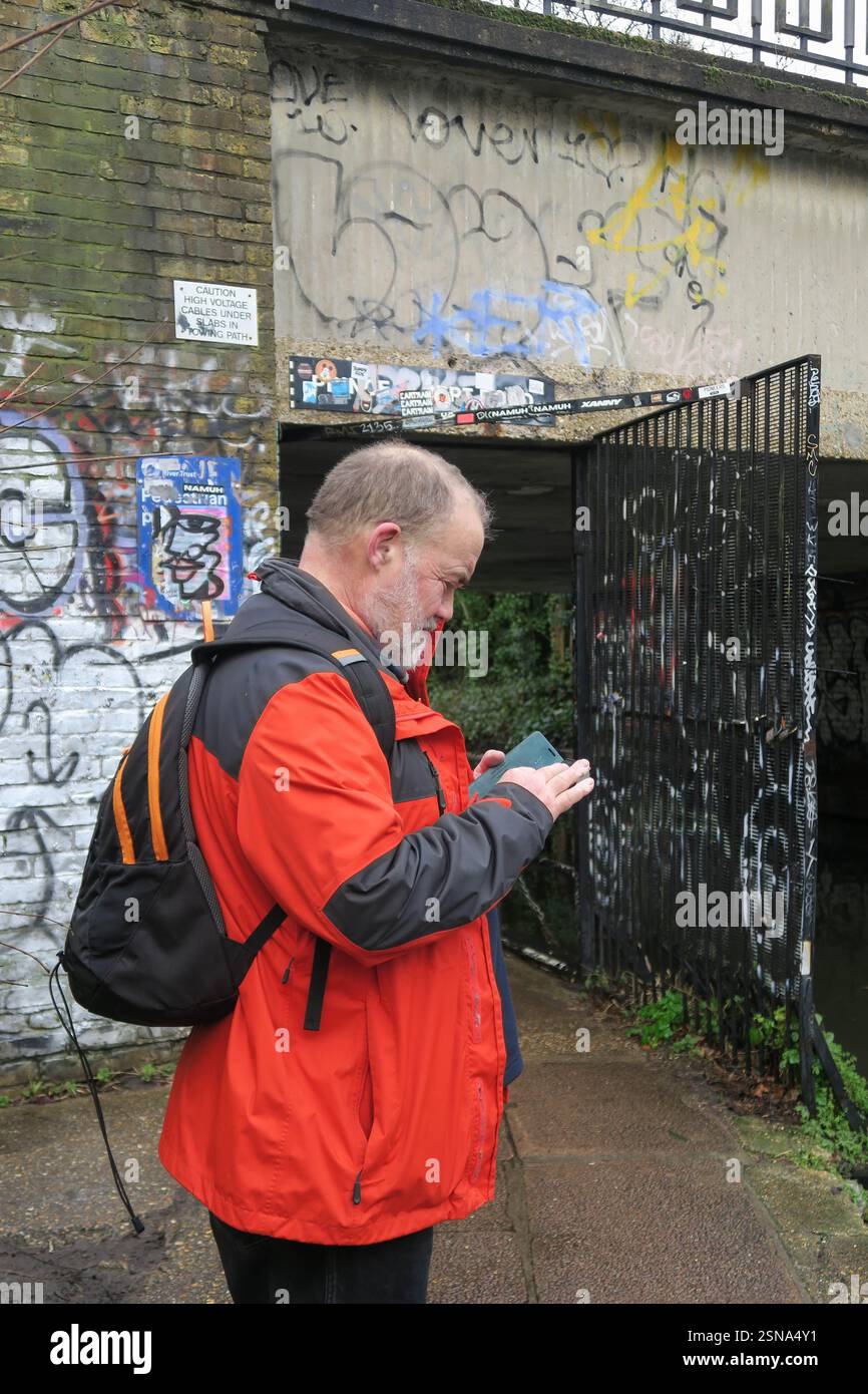 Uomo di mezza età che controlla il suo cellulare per le indicazioni, mentre si trova accanto a un ponte coperto di graffiti sul sentiero del Regents Canal a Londra Foto Stock