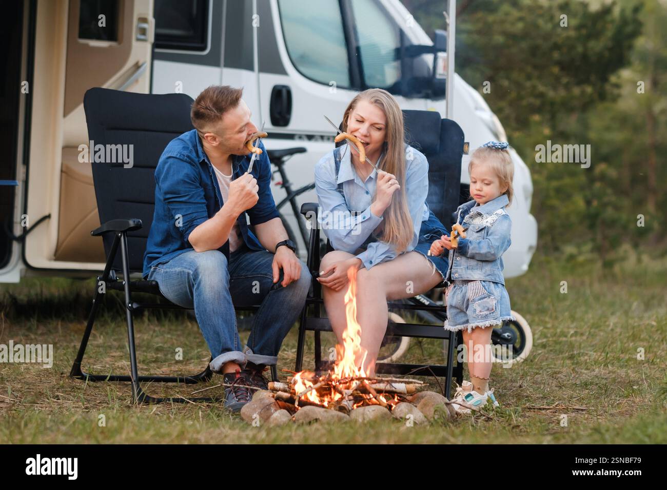 Una famiglia si siede accanto a un falò, arrostendo marshmallow e condividendo sorrisi. Il padre e la madre si divertono mentre la loro giovane figlia si unisce alla f Foto Stock