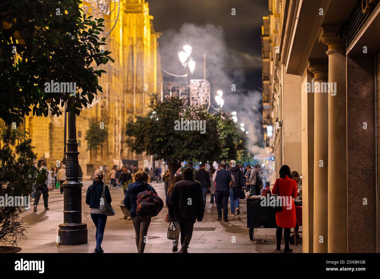 Una donna in abito rosso acquista castagne arrostite durante le festività natalizie accanto alla cattedrale illuminata di Siviglia nel centro di Siviglia in Spagna. Foto Stock