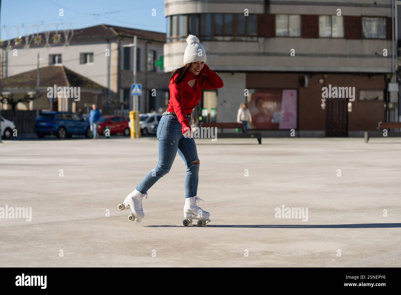 Giovane donna con maglione rosso e pattinaggio a rotelle in jeans gioiosamente in una piazza urbana, abbracciando libertà e movimento in una giornata di sole Foto Stock