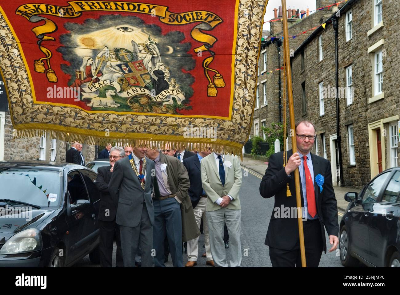 La parata annuale della Askrigg Friendly Society arriva alla chiesa del villaggio dietro lo striscione della Friendly Society "Pace e abbondanza". Fuori dal Kings Arms Hotel, il loro tradizionale luogo di incontro, i membri onorari indossano rosette bianche mentre i membri ordinari indossano blu. Askrigg, North Yorkshire, Inghilterra 5 giugno 2014 2010s UK HOMER SYKES Foto Stock
