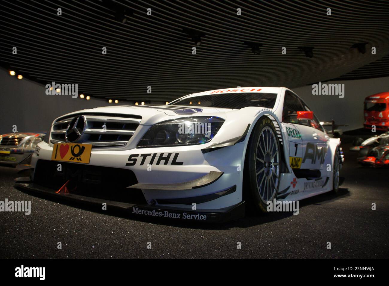 Un'auto da corsa Mercedes-Benz Classe C DTM bianca con accenti rossi è parcheggiata in un museo, circondata da altri veicoli. Questa macchina ad alte prestazioni è una Foto Stock