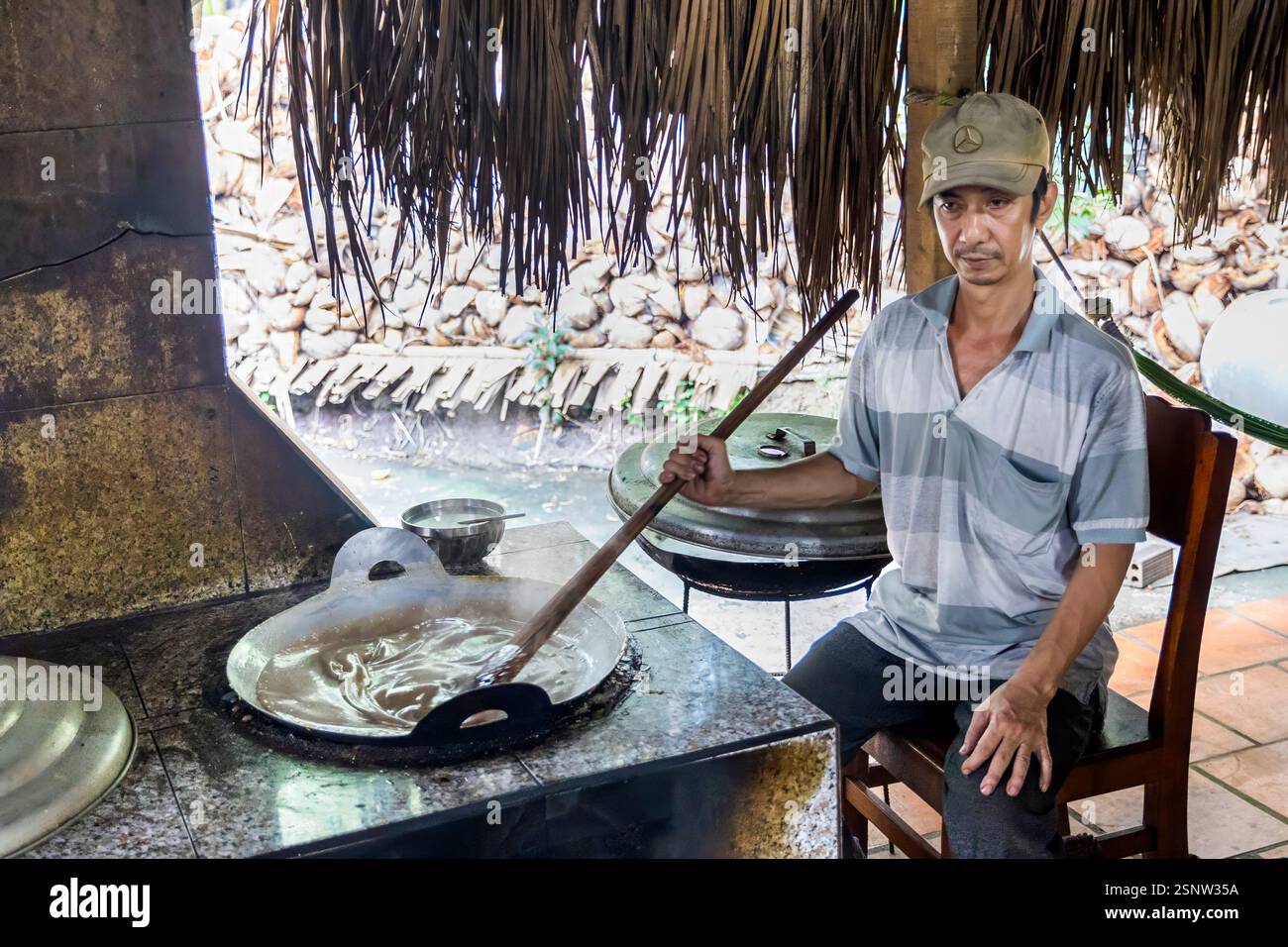 Un cuoco mescola una grande padella in una cucina all'aperto in una fabbrica locale di caramelle al cocco, Quoi Son, Vietnam. Lunedì 11 novembre 2024. Foto Stock