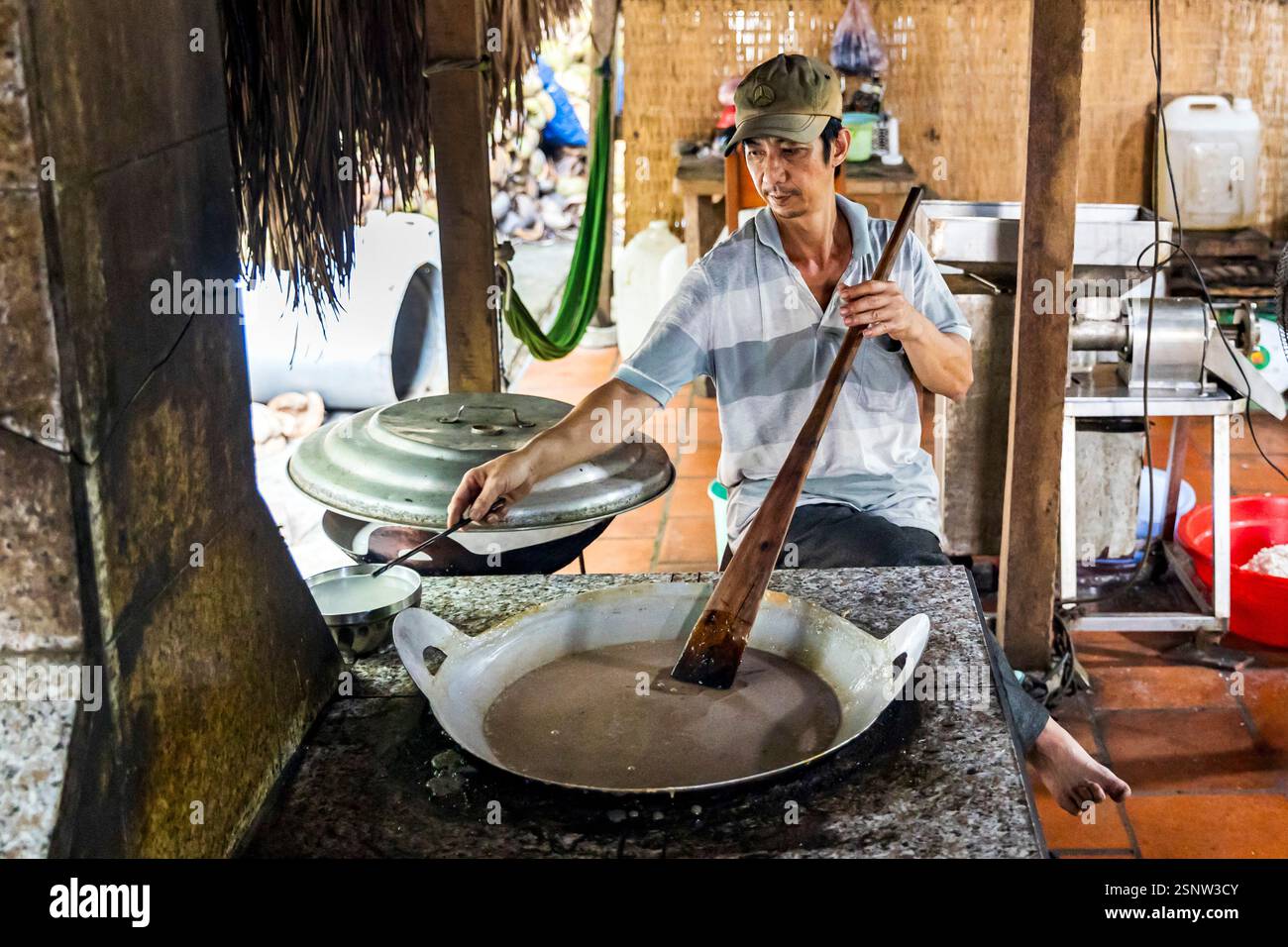 Un cuoco mescola una grande padella in una cucina all'aperto in una fabbrica locale di caramelle al cocco, Quoi Son, Vietnam. Lunedì 11 novembre 2024. Foto Stock