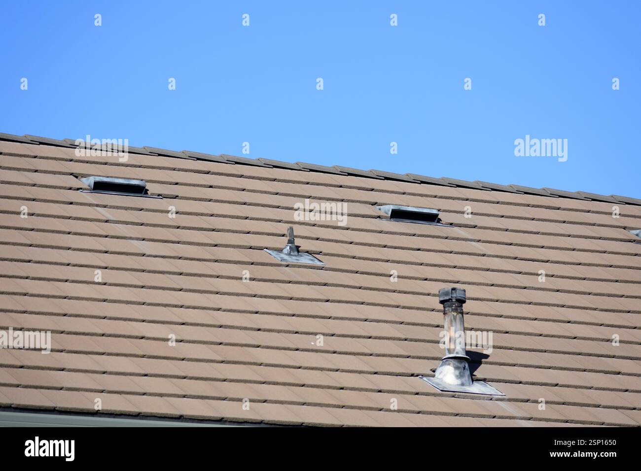 Vista ravvicinata di un tetto inclinato con piastrelle marroni, con due aperture di ventilazione e un tubo di camino in metallo intemprato sotto un cielo azzurro Foto Stock