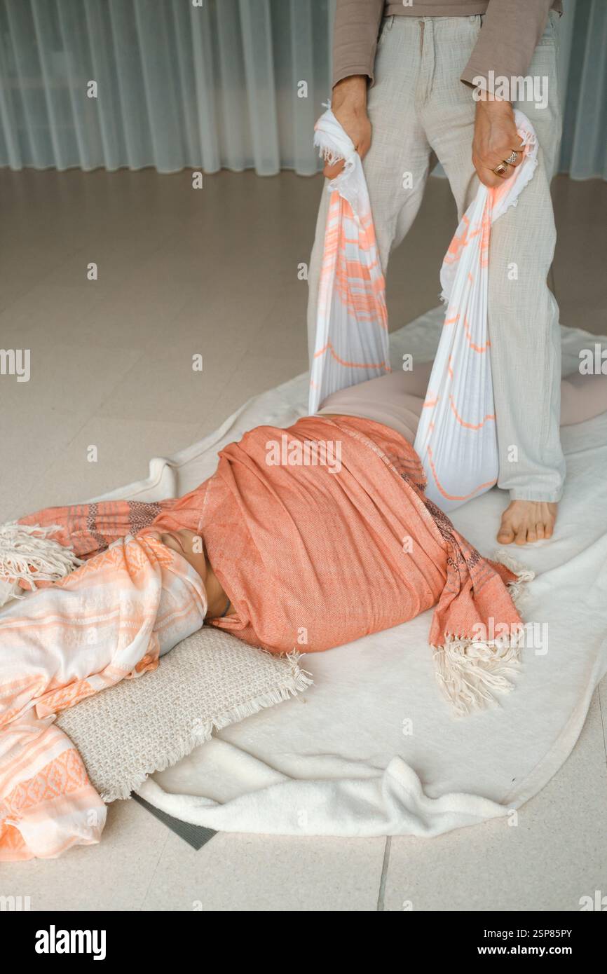 Un praticante assiste delicatamente un partecipante avvolto in un panno morbido sul pavimento di un tranquillo studio, promuovendo il relax attraverso lo stretching guidato e. Foto Stock