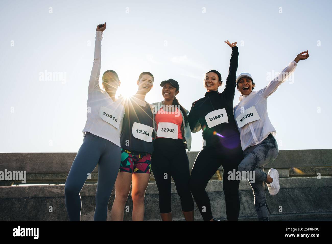 Un gruppo di diverse amiche festeggia una gara su strada. Sono in piedi insieme a pettorali da corsa, braccia sollevate vittoriosamente, catturando lo spirito di f Foto Stock