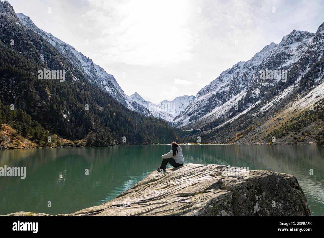 Una donna solitaria siede su una grande roccia, guardando le tranquille acque turchesi del lago di Gaube, circondato da montagne innevate e densa sempreverde Foto Stock
