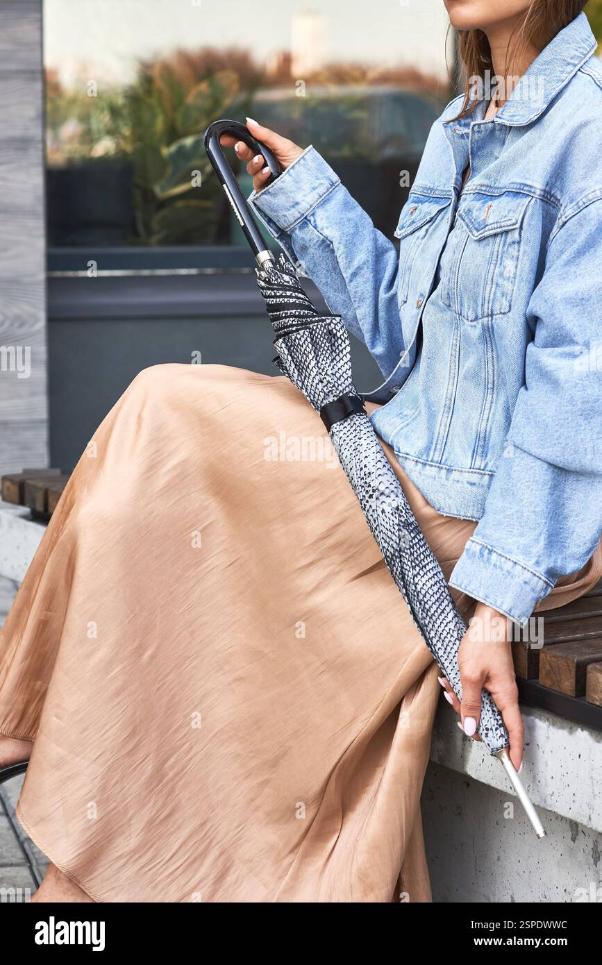 Donna casual con giacca e gonna in denim seduta sulla panca che regge un ombrello chiuso a motivi geometrici, un concetto di stile alla moda Foto Stock
