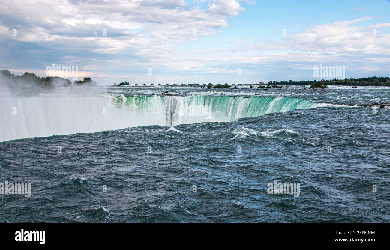 Le potenti acque delle Cascate del Niagara scorrono drammaticamente oltre il bordo sotto un cielo luminoso, circondato dalla natura. Foto Stock