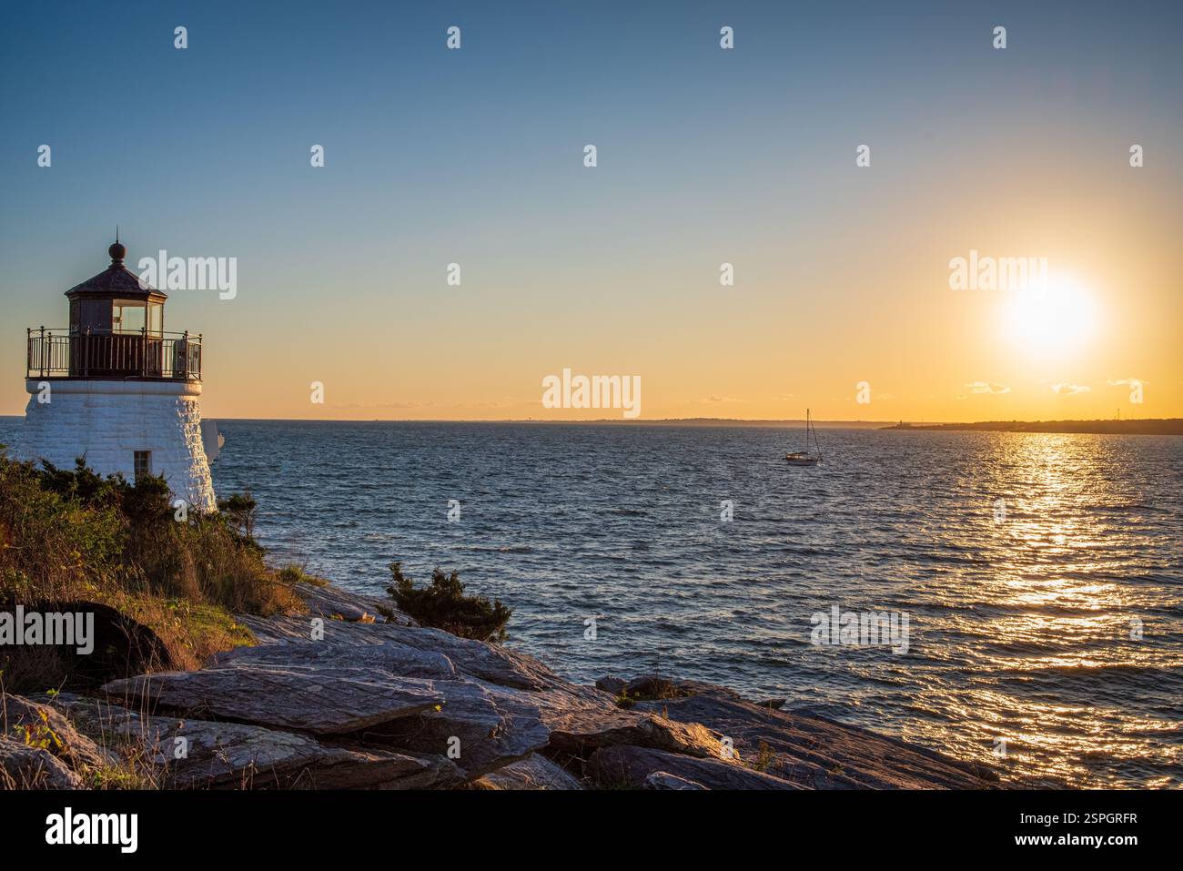 Un faro su una costa rocciosa si affaccia su una barca a vela che naviga sull'oceano mentre il sole tramonta, gettando luce dorata sull'acqua. Foto Stock