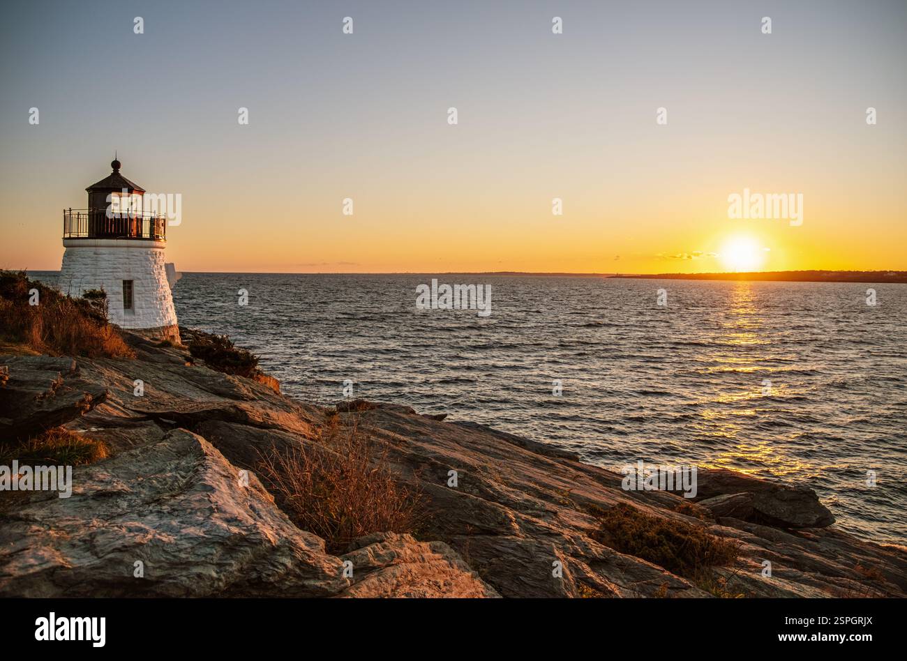Uno storico faro sorge su una costa rocciosa, affacciato sull'oceano mentre il sole tramonta, che getta luce dorata sull'acqua nel Rhode Island. Foto Stock