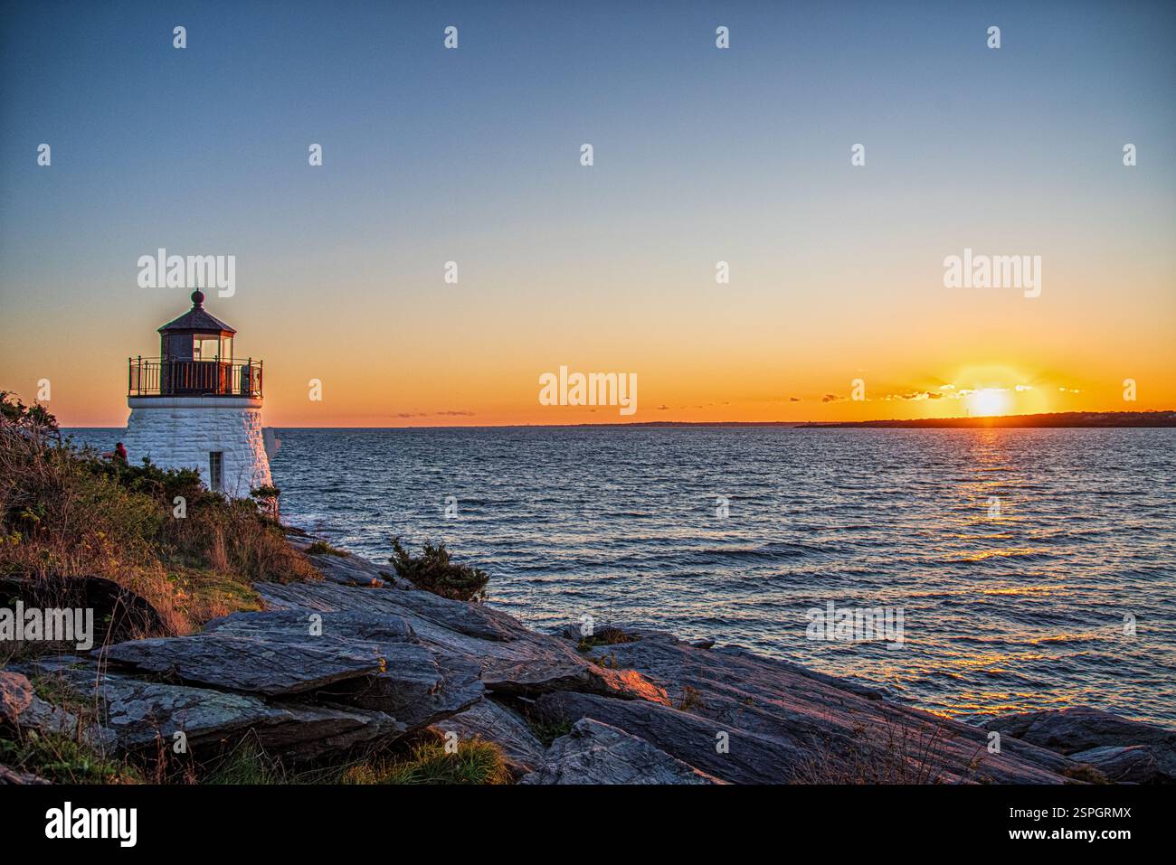 Un faro costiero sorge su una costa rocciosa mentre il sole tramonta sull'oceano, proiettando un bagliore dorato sull'acqua e sul cielo. Foto Stock