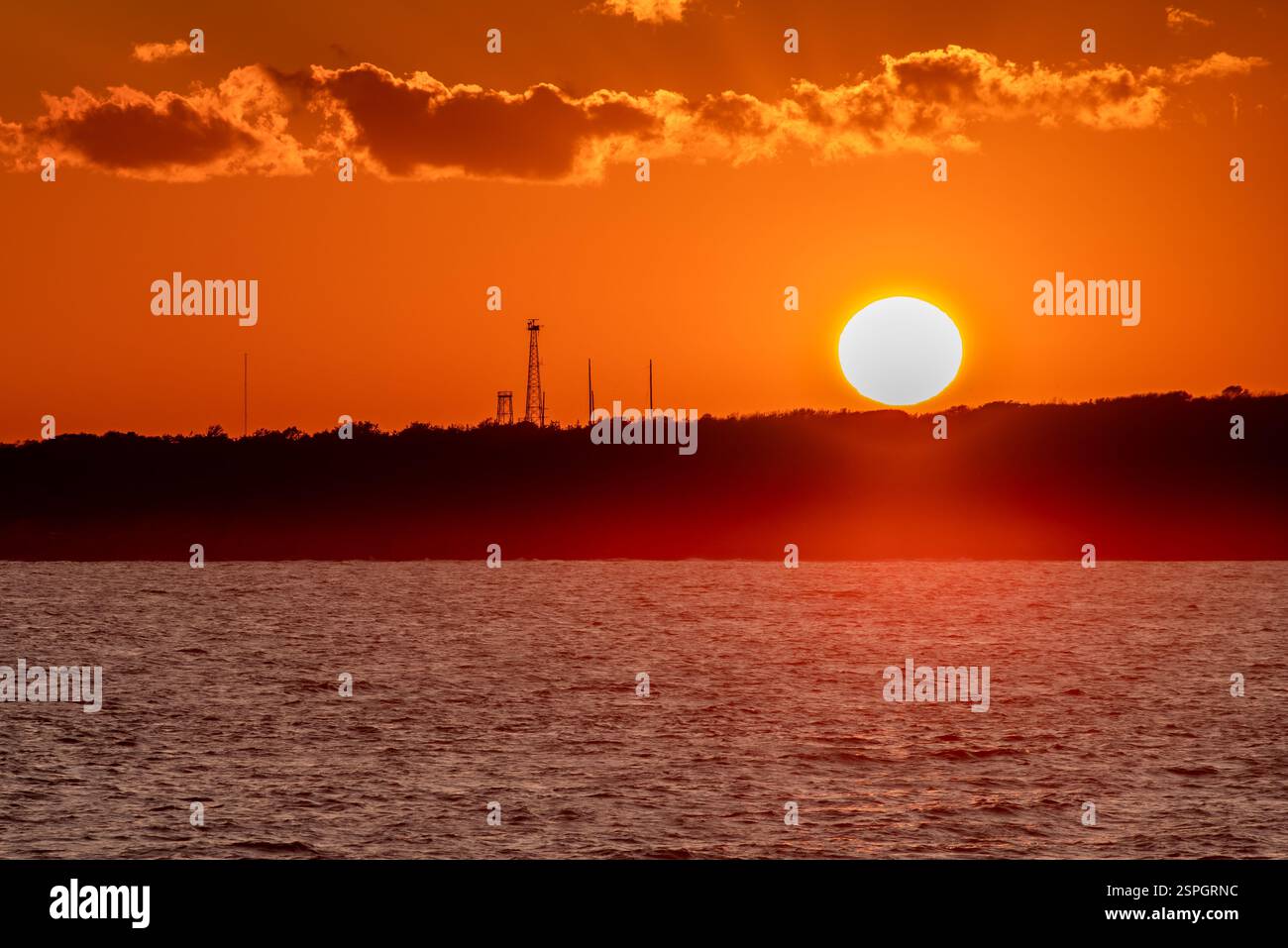 Un drammatico tramonto rosso e arancione sull'oceano, con strutture industriali sagomate lungo l'orizzonte. Foto Stock