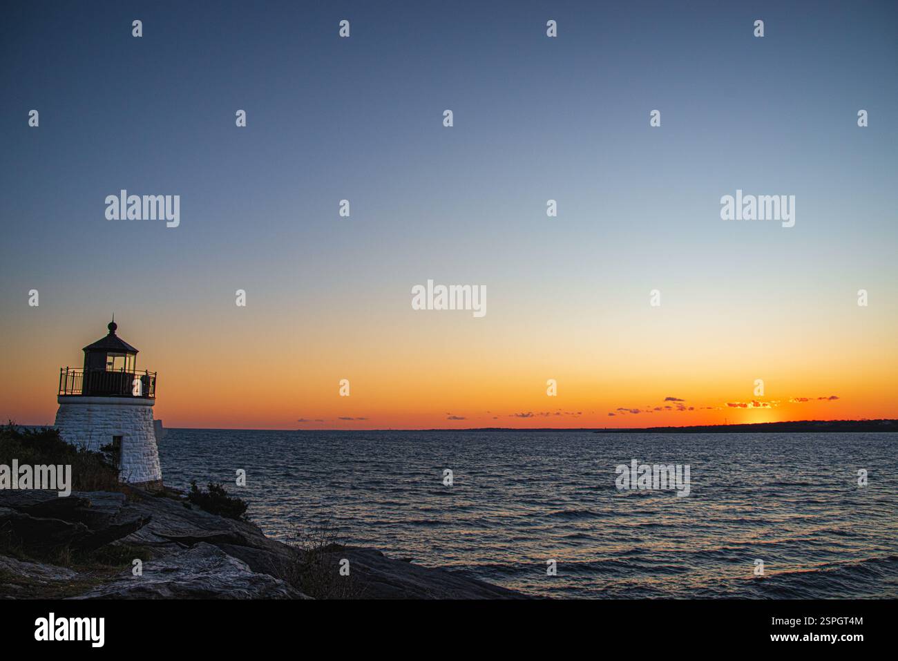 Un faro panoramico sorge sulla costa rocciosa mentre il sole tramonta sull'oceano, proiettando calde sfumature attraverso il cielo e riflettendo sull'acqua. Foto Stock