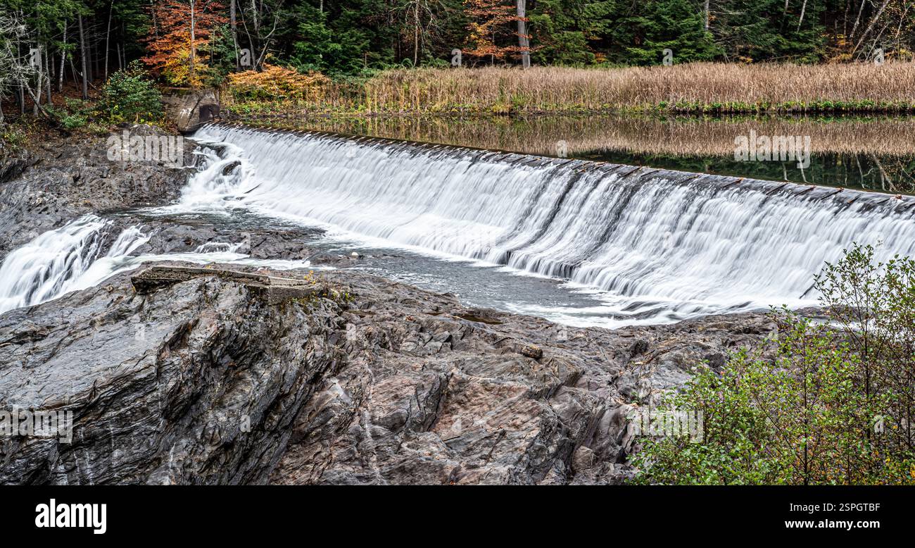 Cascata su una diga artificiale circondata da fogliame autunnale e rocce aspre, che riflettono la bellezza stagionale e l'idroingegneria. Foto Stock