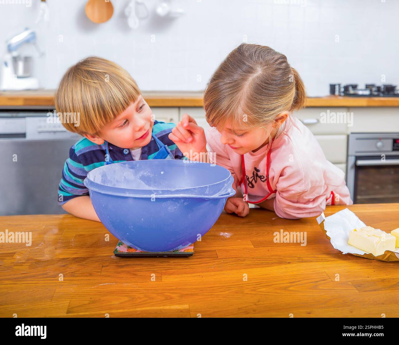 Due bambini amano cucinare in una cucina moderna e luminosa, misurando gli ingredienti e condividendo momenti di gioia. Foto Stock