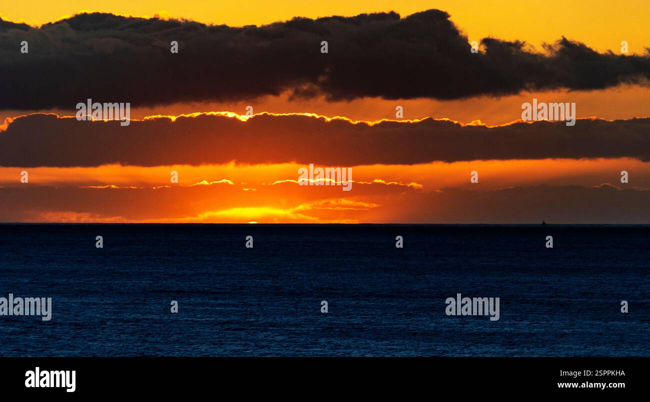 Uno splendido tramonto sull'oceano, con vivaci sfumature arancioni e gialle che illuminano l'orizzonte. Le nuvole scure incorniciano la scena, creando un contorno drammatico Foto Stock