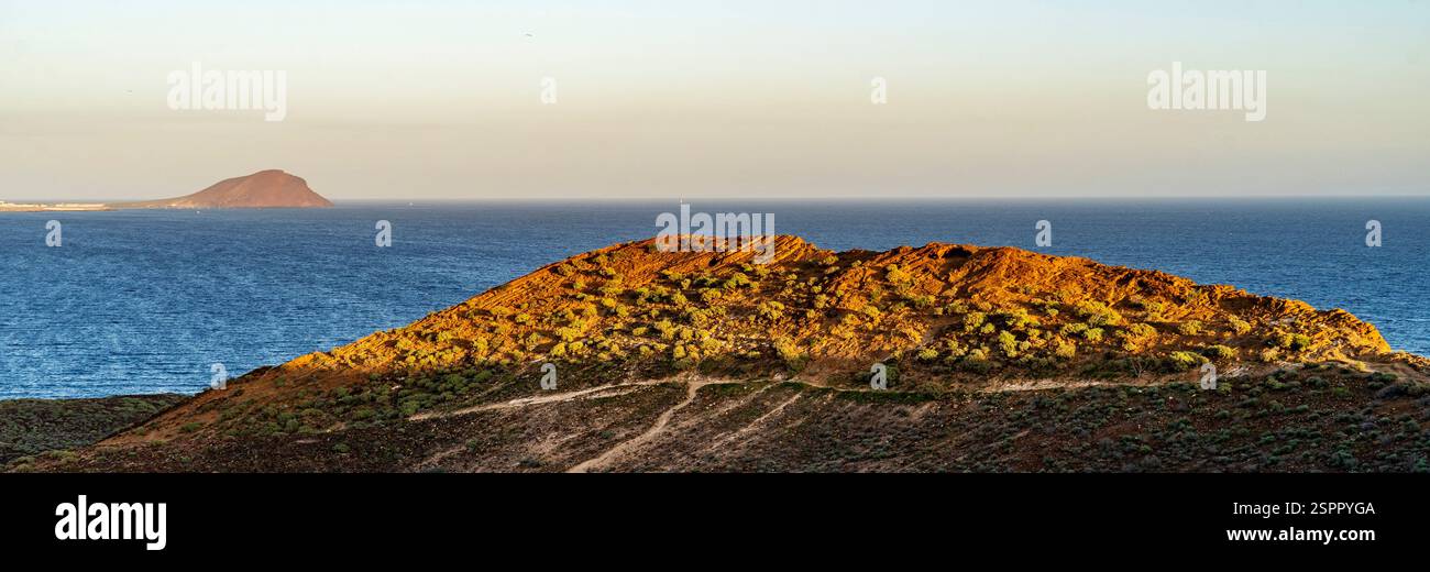 Una vista panoramica di un paesaggio costiero caratterizzato da una collina rocciosa in primo piano e da un'isola lontana all'orizzonte. Il sole proietta un caldo bagliore sul ciglio Foto Stock