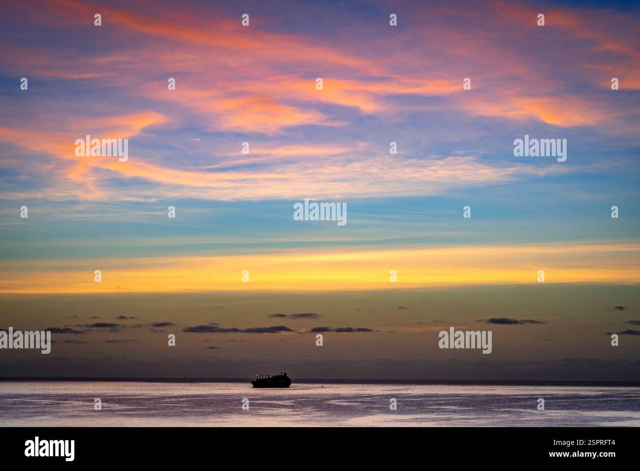 Un tranquillo paesaggio marino al tramonto, caratterizzato da un oceano calmo che riflette vibranti colori del cielo. Una sagoma di una nave è visibile all'orizzonte contro una B. Foto Stock