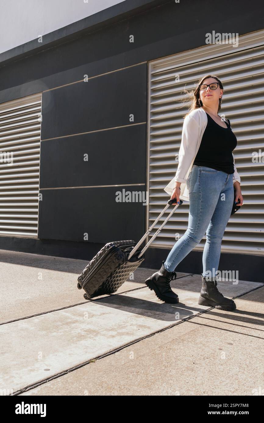 Giovane donna d'affari che tira la valigia mentre cammina fuori dal terminal dell'aeroporto, si prepara a imbarcarsi in un viaggio di lavoro, rappresentando viaggi, autobus Foto Stock