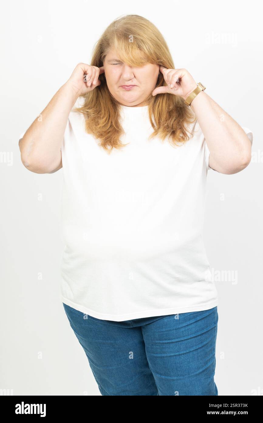 Ritratto di una donna adulta che copre le orecchie con le dita e chiude gli occhi con frustrazione. Lo sfondo bianco enfatizza il suo corpo Foto Stock