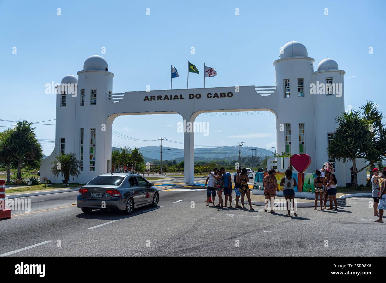 Vista completa del cancello della città di Arraial do Cabo con turisti, un'auto di passaggio e bandiere ondulate sotto un vivace cielo azzurro e soleggiato al mattino d'estate. Foto Stock