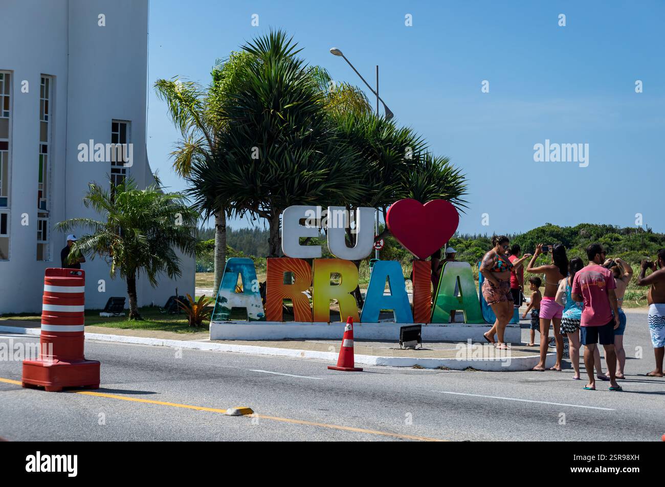 I turisti che scattano foto al vibrante cartello della città "i Love Arraial do Cabo" circondato da alberi sotto il sole del mattino d'estate, il cielo azzurro e limpido. Foto Stock