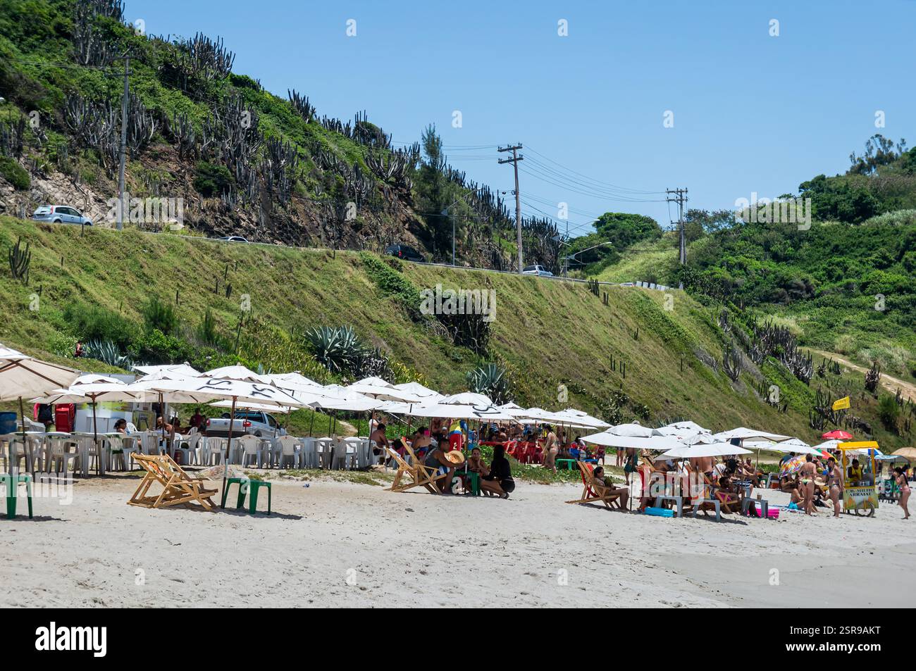 Estremità nord-occidentale della spiaggia di Prainha con sedie a sdraio e ombrelloni, un carrello per il cibo e una collina verde sul retro sotto il cielo azzurro e soleggiato. Foto Stock