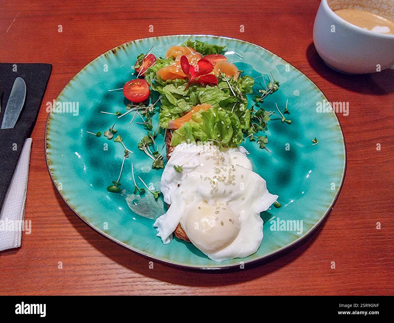 Uovo in camicia con verdure fresche (verdure, lattuga, pomodoro, crescione) e un fiore commestibile rosso decorativo su un piatto e cappuccino a colazione o a pranzo - Immagine stock catturata con smartphone