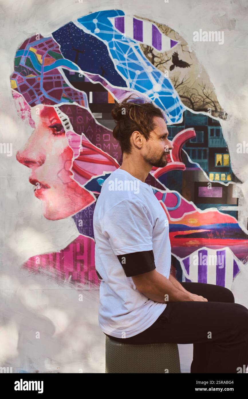 Giovane maschio adulto con capelli castani seduto, contemplativo, prima di un murale colorato. Design luminoso e astratto in background, che unisce tecnologia e natura Foto Stock