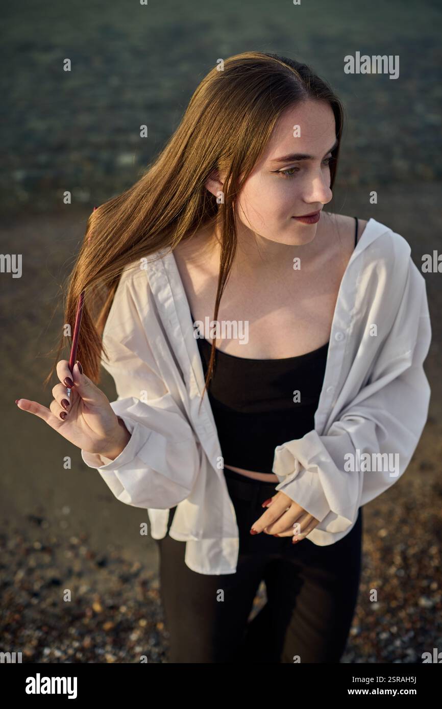 Una giovane donna si staglia serenamente lungo il litorale, drappeggiata in camicia bianca su un outfit nero. Il sole tramonta diffonde una calda luce soffusa, migliorando la calma e l'atmosfera riflessiva di Foto Stock