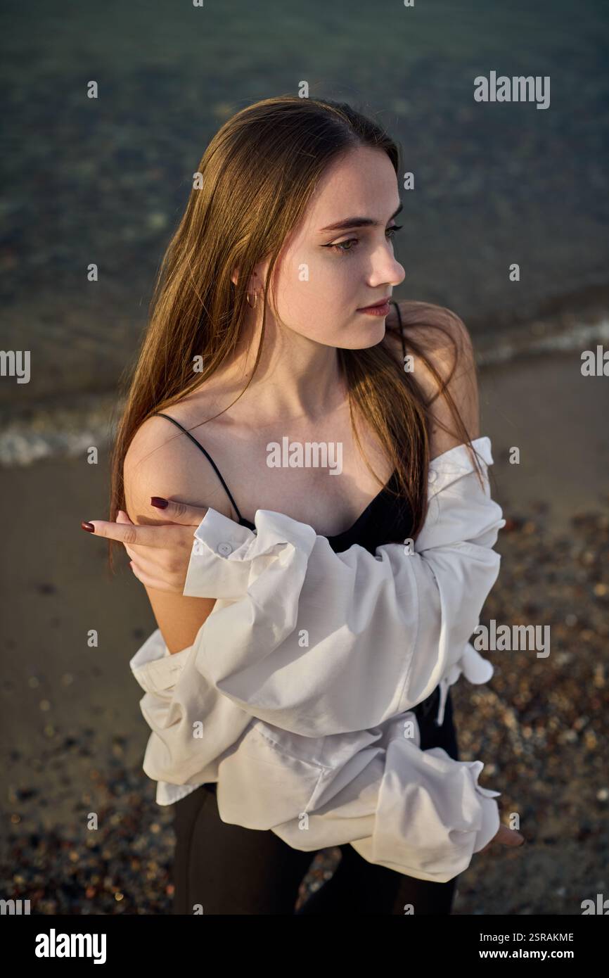 Una giovane donna si staglia serenamente lungo il litorale, drappeggiata in camicia bianca su un outfit nero. Il sole tramonta diffonde una calda luce soffusa, migliorando la calma e l'atmosfera riflessiva di Foto Stock
